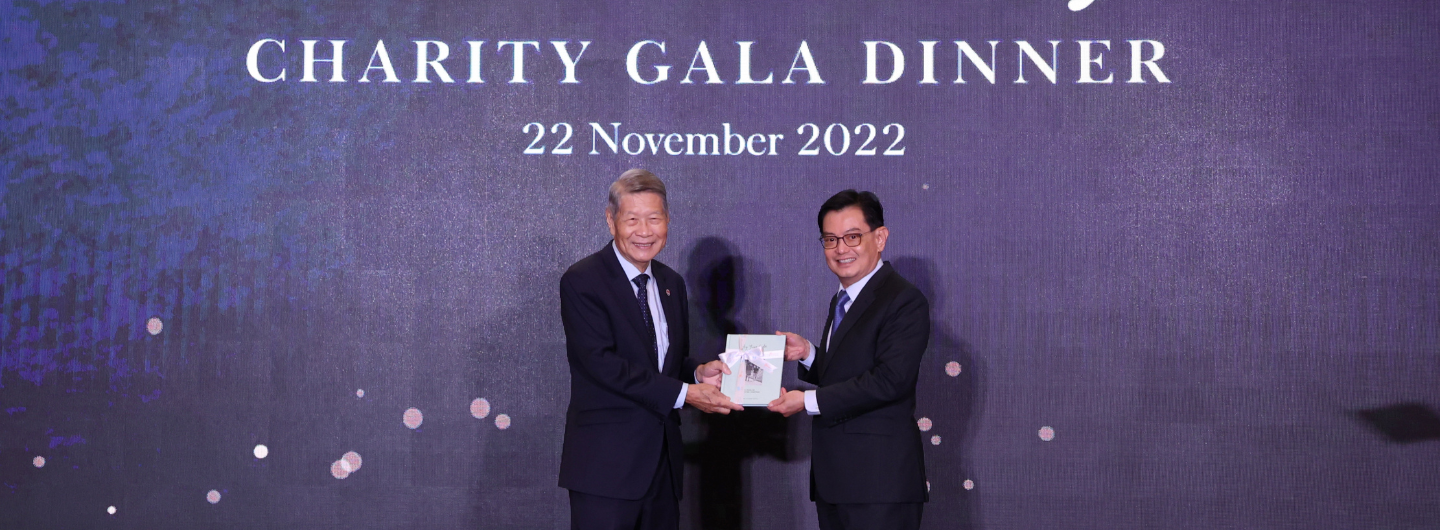 Two men in suits exchange a book in front of a "Charity Gala Dinner 22 November 2022" backdrop.